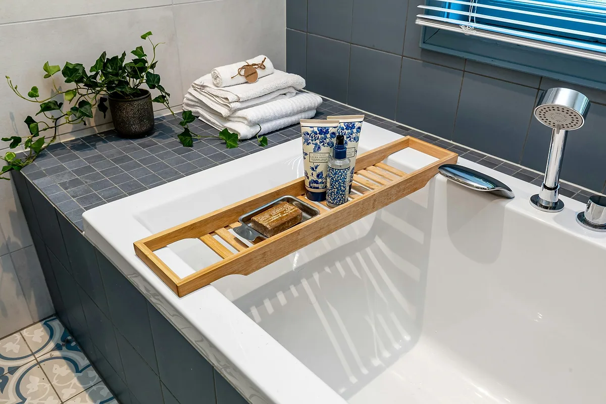 Bamboo bathtub tray across a white tub holding soap and bath bottles, with folded towels and ivy on the tiled ledge