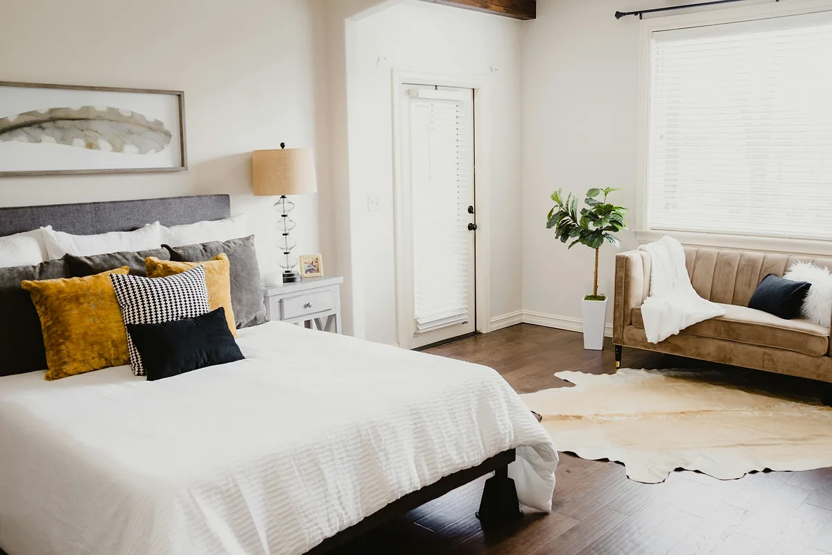 Cozy bedroom with white bedding, mustard and black throw pillows and a small sofa by the window