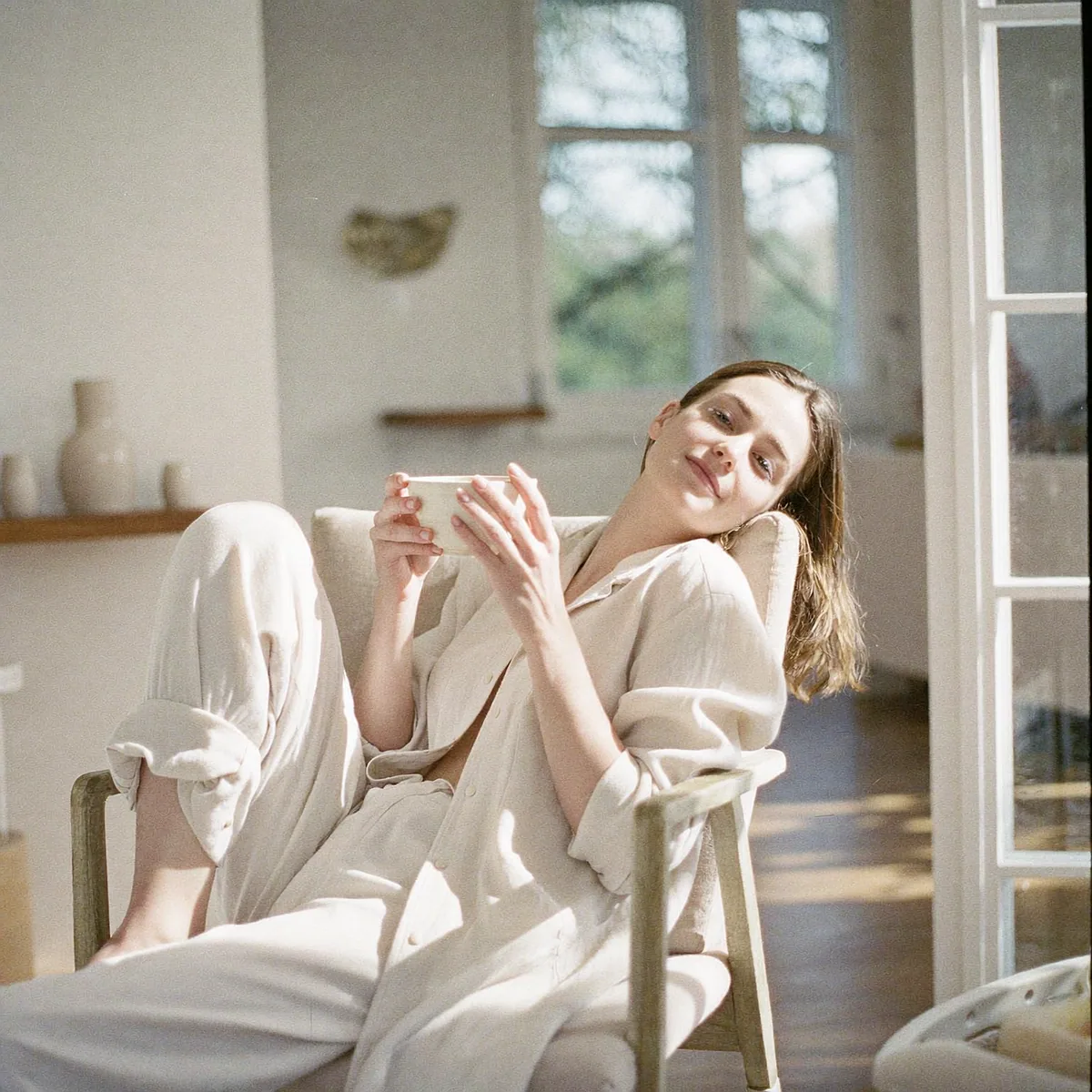 Woman in a white linen outfit relaxing in an armchair with a cup in a sunlit room